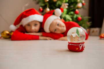 two children, brother and sister in red Christmas pajamas look at a glass ball with a Christmas tree and a house inside, dream and make wishes. new Year's winter concept. celebrating at home