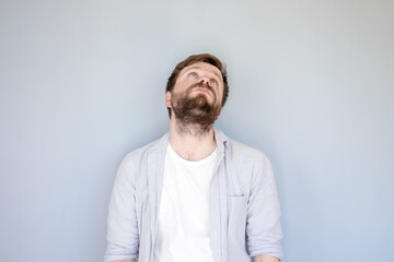 Unhappy, tired Caucasian bearded man in a shirt raised head and looked up, hands down in hopelessness. Gray background.