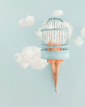 Cute beautiful female doll with head in clouds trapped in a blue cage hovering in space against pastel blue background. Minimal creative concept. Freedom, daydreaming and imagination note card idea.
