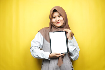 Beautiful young asian muslim woman smiling, excited and cheerful holding tablet with white/blank screen, promoting app, promoting product, presenting something, isolated on yellow background