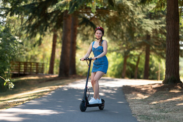 An active girl in a safety helmet and a denim dress rides an electric scooter. Beautiful nature, sunny day. Eco-friendly transport. Color image.