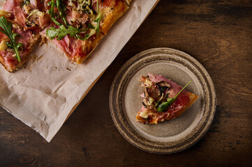Piece of Italian Roman pizza lies on rustic ceramic plate on brown wooden background, next to pizza on craft paper, top view