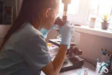 Asian woman lab assistantlooks into microscope at sample sitting near window in hospital