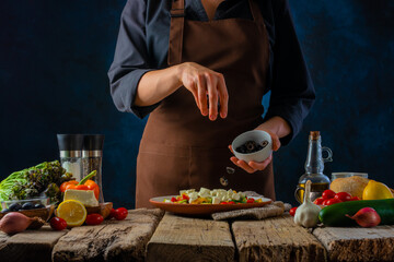 The chef adds the olives to the bowl. Lots of ingredients. Cooking Vegetarian Greek Salad. Wooden texture. Culinary blog, recipe book, menu design. Close-up.