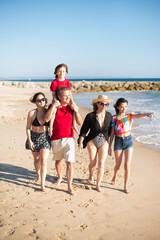 Cheerful family at sea coast. Children and parents walking by sea, smiling, boy sitting at shoulders, girl pointing. Family, vacation, outdoor activity concept