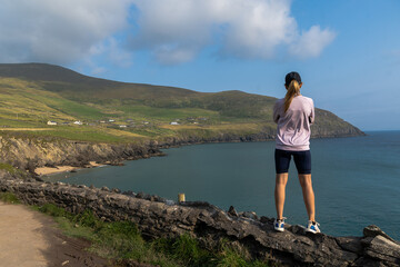 Shot of a young Caucasian woman standing on a wall looking at cliffs on Dingle peninsula, Ireland