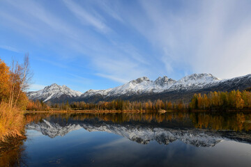 Fototapeta na wymiar Fall colors, snowy mountains and fluffy clouds reflect in the waters of Alaska's Reflections Lake.