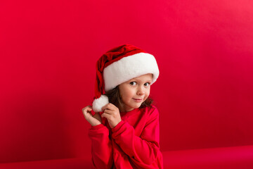 Cute little girl in a red Christmas hat and red dress on a red background. Merry Christmas and Happy Holidays.	