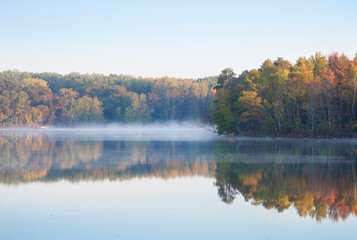 Fototapeta premium Trees in autumn color on a misty calm lake in northern Minnesota at dawn