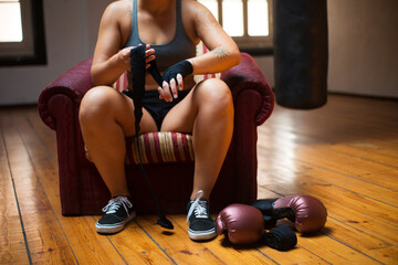 Close-up of young woman applying hand wrap. Sporty woman in black sport clothes sitting in chair,...
