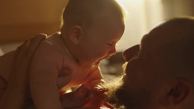 Young father with beard caressing and kissing his newborn baby kept by mother in arms in kitchen at home in the morning. Concept of family, fatherhood, motherhood, parenthood, childhood, life.