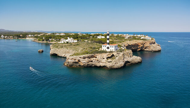 The lighthouse of Porto Colom on the Spanish Mediterranean island of Mallorca in sunshine in summer. A boat drives towards the harbor. In the foreground are the cliffs.