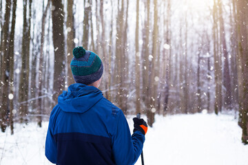 Skier in windbreaker and hat with pompom with ski poles in his hands with his back against the background of a snowy forest. Cross-country skiing in winter forest, outdoor sports, healthy lifestyle. © Ольга Симонова