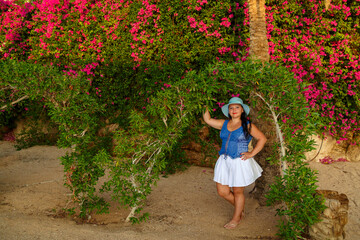 A brunette woman in a white skirt and a sun hat stands by the flowering plants.