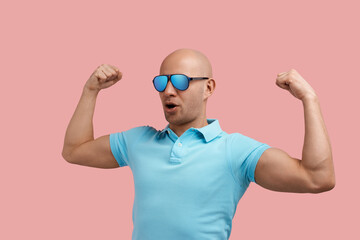 Fototapeta premium Fitness addicted strong bald homosexual man with bristle shows muscles, biceps with both hands, hits gym, lives healthy, gay friendly, wears blue polo shirt and sunglasses, stands on pink background.