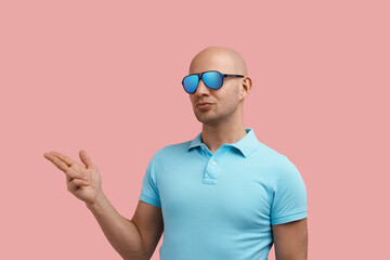 Naklejka premium Handsome homosexual man with bristle puts lips, makes shooting gesture with fingers aside, advertises secret deal, gay friendly, wears blue polo shirt and sunglasses, stands on pink background.