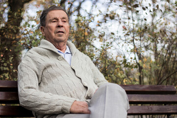 Fototapeta premium Portrait of a pensive senior man sitting on the bench, in the public park, outdoors. Old man relaxing outdoors and looking away. Portrait of elderly man enjoying retirement