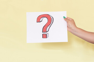Latin woman's hand holding a sign with a question mark in red on a white sheet, on yellow background, unrecognizable model.