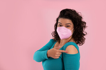 .young latin woman wearing protective mask and pointing arm, vaccine site, on pink background.