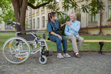 Full length shot of young nurse wearing face shield supporting aged man, recovering male patient, sitting together on the bench in park near hospital