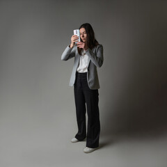 businesswoman dressed in a jacket and trousers in a casual style uses smartphone for work and contact with partners, studio portrait on gray background, Use touch screen, take photo or use video call