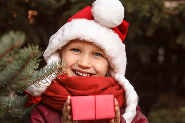 Smiling child in a Christmas hat holds a gift box near the Christmas tree