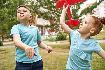 Obraz premium Little girl and boy with airplane toys. Adventure fly concept. Stay home game. Family garden activity. Red hair female portrait. Styrofoam childhood pilot. Summer time. Park outdoor aviation. Siblings