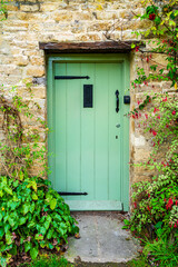 Fototapeta na wymiar Wooden door entrance into rural cottage house in Bibury, Cotswolds, Egland, UK. Vintage wooden door on stone wall with green bushes