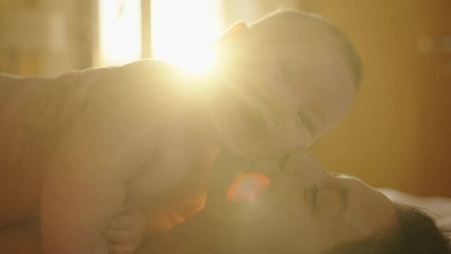 asian woman mother kissing baby and holding infant in arm playing and talking with kid on sofa couch in living room at home in evening. happy baby learning communication by smile, laugh, and Stick out