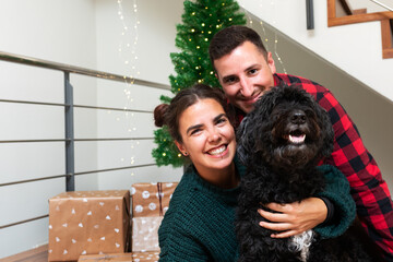 Happy caucasian couple with their black poodle dog looking at camera. Christmas tree and present on...