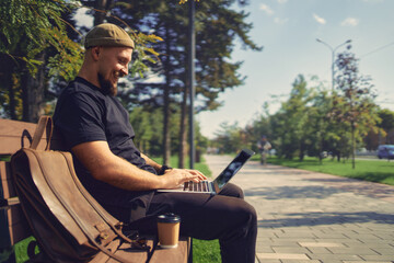 Positive man with laptop doing remote work outside while sits on bench in city park. Distance Job 