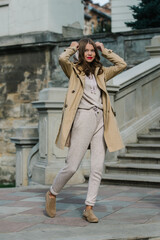 Portrait of fashionable women in beige sports suit, trench coat and stylish suede loafer posing on the stone stairs
