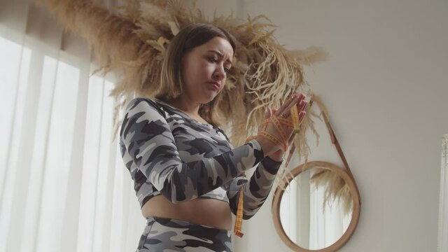 Portrait of unhappy pretty sporty fit woman with hands tied with measuring tape trying to free of constant worry and obsession of weight gaining , expressing determination in domestic room.