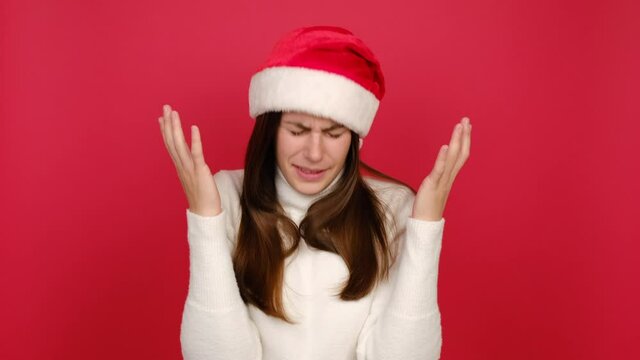 Portrait of sad upset young woman looking camera crying waving hands ask why sadness problem, wears white sweater and Santa Christmas hat, isolated on red color background studio. New Year concept