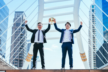 Successful businessman celebrating his victory with arms up and holding gold trophy on cityscape...