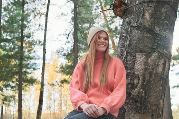 young red haired woman wearing orange pullover sitting in a forest under the tree and smiling. front view. autumn in a forest, warm fall season walking