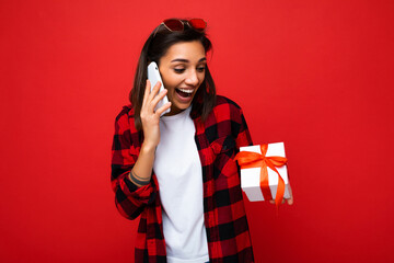 Photo shot of pretty positive surprised young brunet woman isolated over colourful background wall wearing trendy outfit look holding gift box and looking at present box with red ribbon