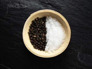Coarse sea salt and whole black peppercorns in bamboo bowl on slate worktop, background. Flat lay, top view.