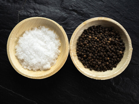 Coarse sea salt and whole black peppercorns in bamboo bowls on slate worktop, background. Flat lay, top view.