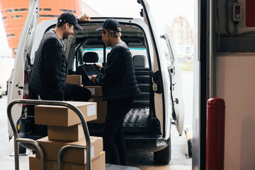 Naklejka premium Back view of two delivery people checking information on a digital tablet while standing at van trunk near a warehouse