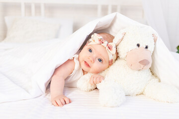 a baby girl lies under a blanket with a teddy bear toy and smiles on a white cotton bed at home