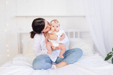 Obraz na płótnie Canvas mom and baby girl hug and kiss sitting on a white cotton bed at home, maternal love and care