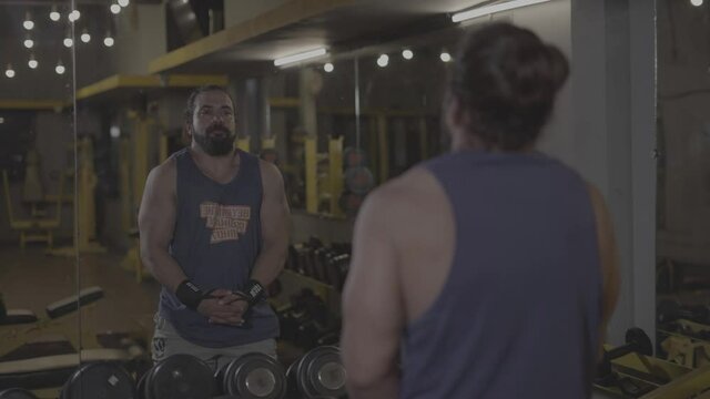 s log. handsome muscular bodybuilder with beard and man-bun doing warm up stretching exercises in front of mirror before workout in gym. fitness workout video