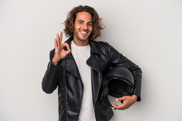Young biker caucasian man holding a motorbike helmet isolated on gray background cheerful and...