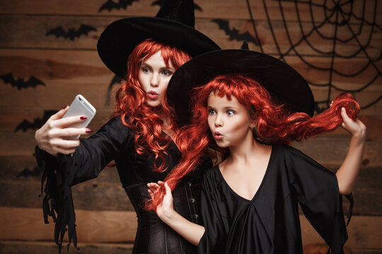 Halloween Concept - Beautiful caucasian mother and her daughter with long red hair in witch costumes taking a selfie with smartphone ready for celebrating Halloween.