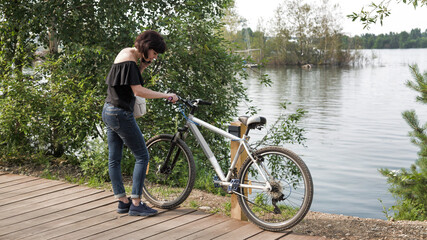 A frustrated woman with a broken bicycle stands by the river calls her husband waiting for help.
