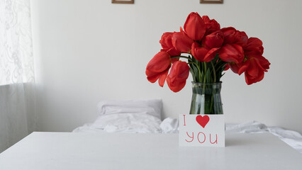 Valentine's day bouquet of red tulips in a glass vase on the table in the bedroom. Postcard with the inscription I love you.