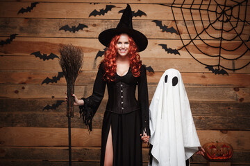 Fototapeta na wymiar Halloween Concept - Witch mother and little white ghost doing trick or treat celebrating Halloween posing with curved pumpkins over bats and spider web on Wooden studio background.