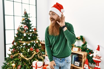 Redhead man with long beard wearing christmas hat by christmas tree smiling with hand over ear listening an hearing to rumor or gossip. deafness concept.