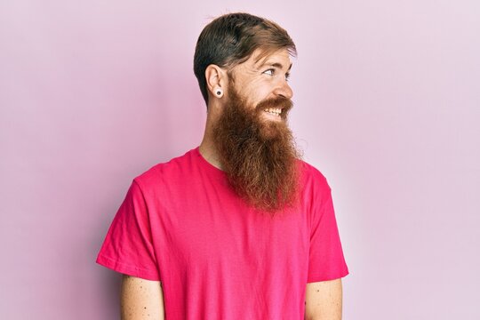 Redhead man with long beard wearing casual pink t shirt looking to side, relax profile pose with natural face and confident smile.